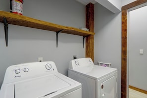A laundry room with two white front-loading washing machines, wooden shelves above, and a wooden column. - Walk to Creek! 10-Acre 'Spring Creek Lodge' (Blue Ridge)