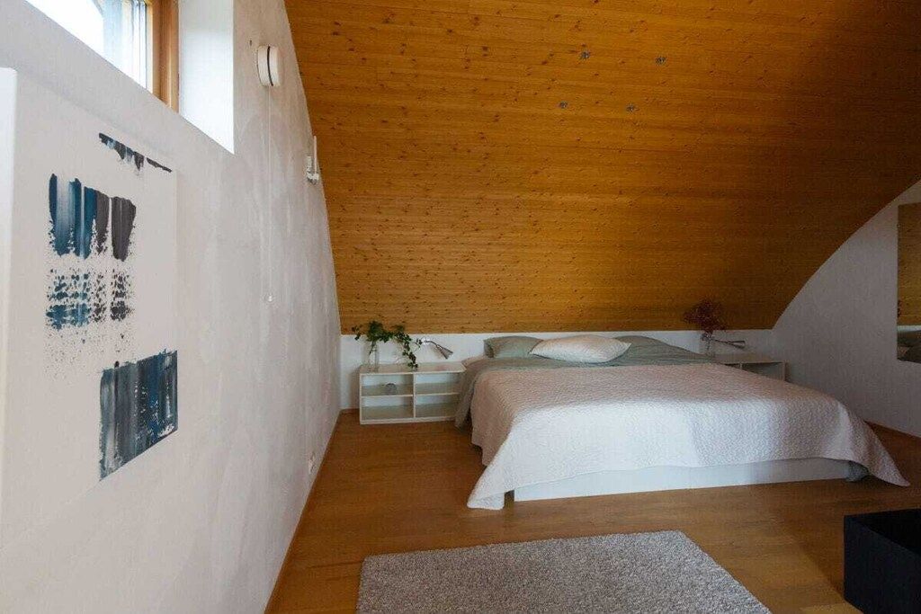 Une chambre avec un plafond en pente, un lit recouvert d’une literie blanche, une table de chevet agrémentée d’une plante, et un mur orné d’œuvres d’art abstraites.