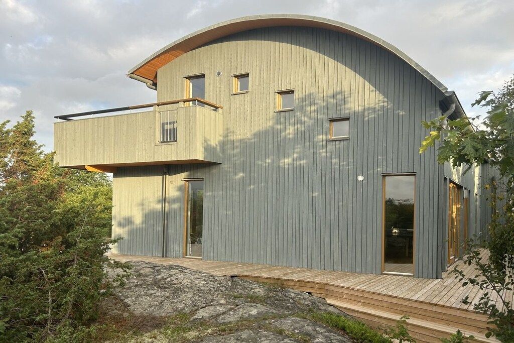 Ein modernes, zweistöckiges Haus mit gewölbtem Dach, einer Holzterrasse und einem großen Fenster.
