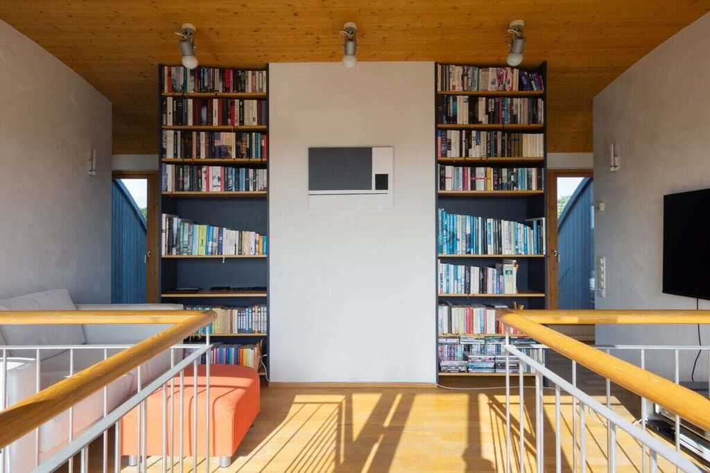 Ein Zimmer mit Holzboden, einer weißen Wand und zwei Bücherregalen voller Bücher. Es gibt ein kleines Fenster und einen an der Wand befestigten Fernseher.