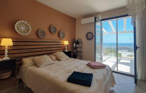 Een slaapkamer met een bed, twee bedlampen, decoratieve wandplaten, een klein plankje en uitzicht op zee vanaf een balkon.