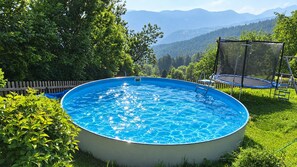 Piscina circular sobre terra amb una escala, envoltada de vegetació abundant i una tanca de fusta, amb vista a muntanyes llunyanes.