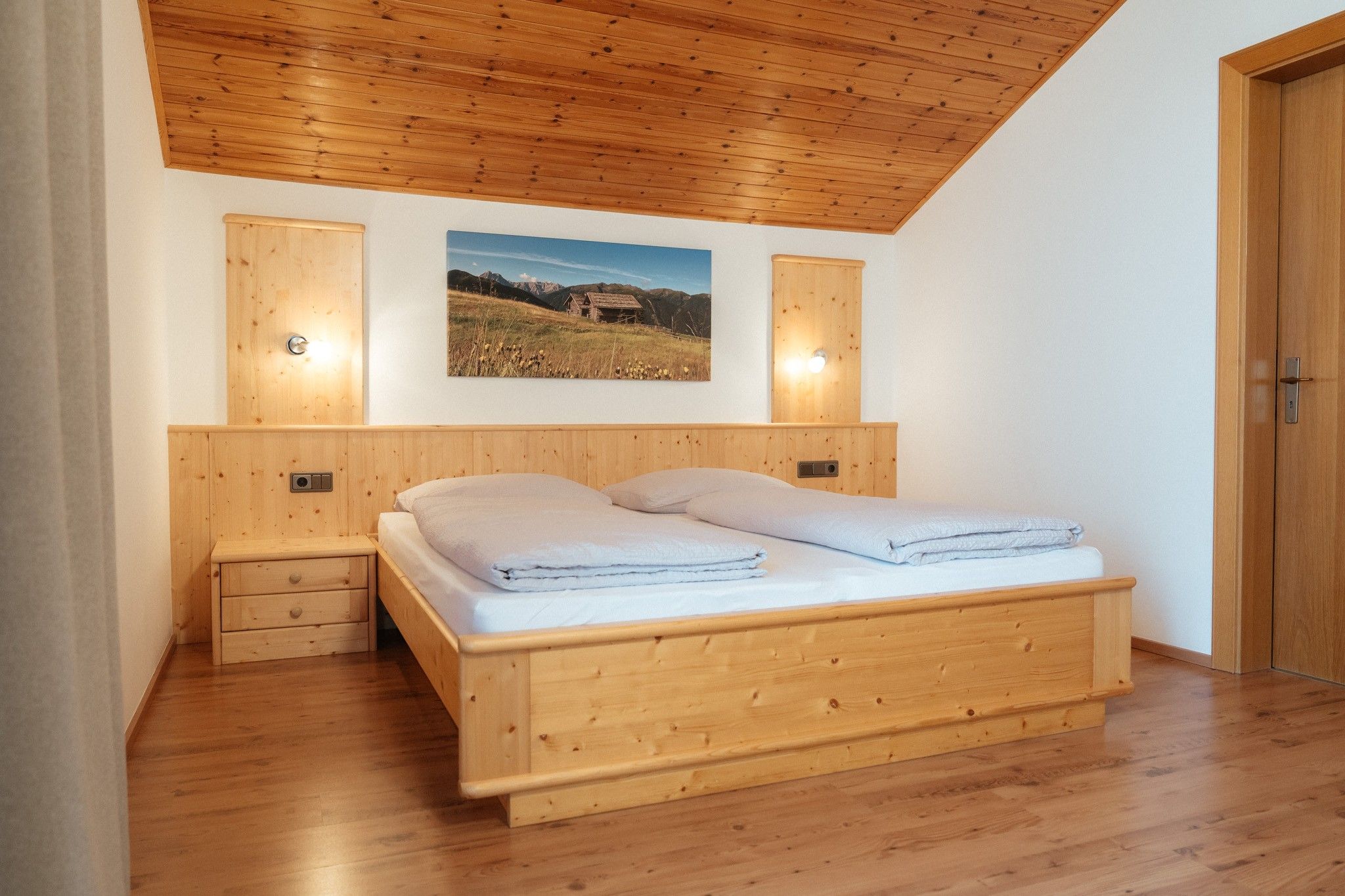 Une chambre avec un plafond en bois, un lit recouvert de draps blancs, une table de chevet et un tableau accroché au mur.