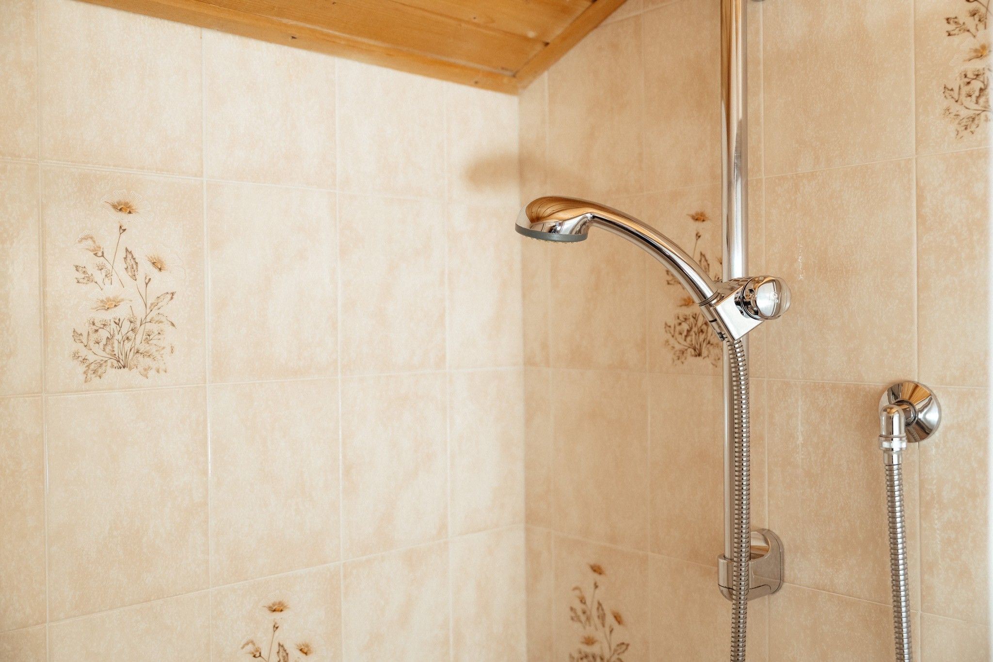 Une douche dotée d’un robinet et d’un flexible chromés, installée sur un mur recouvert de carreaux beiges ornés de motifs floraux.