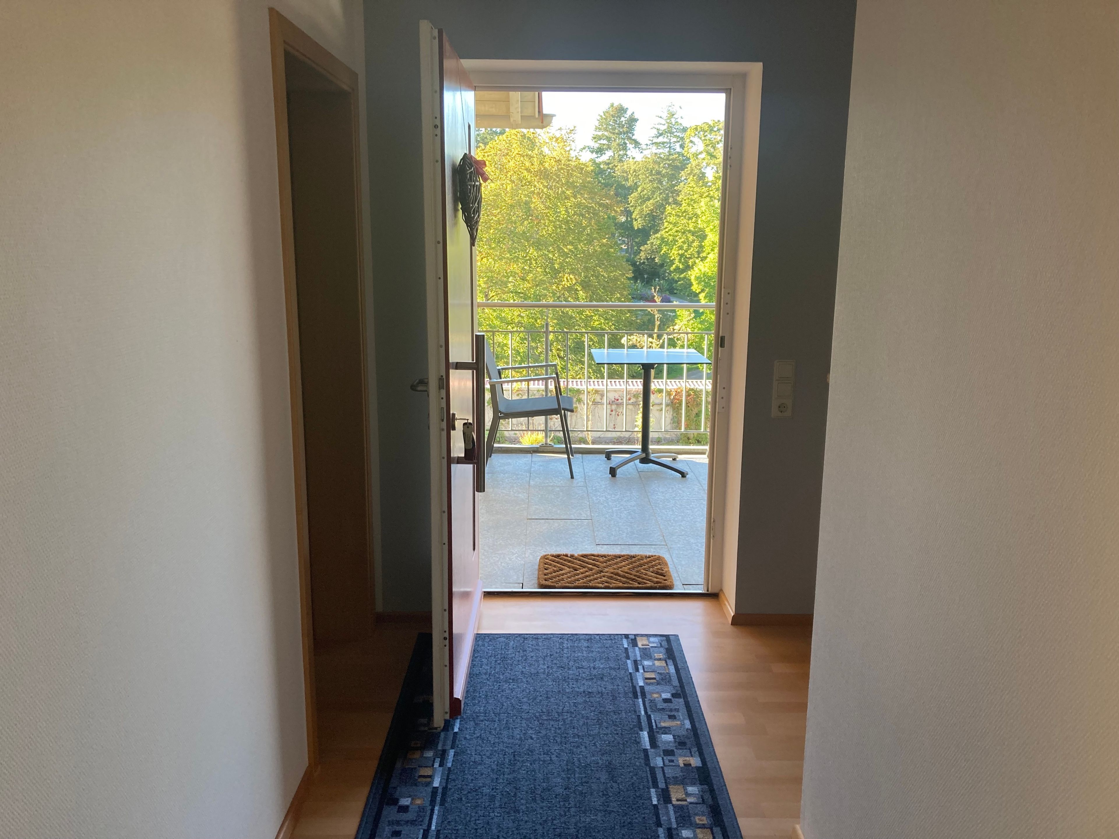 A hallway leading to a balcony with a table and chairs, a doormat, and a view of trees.