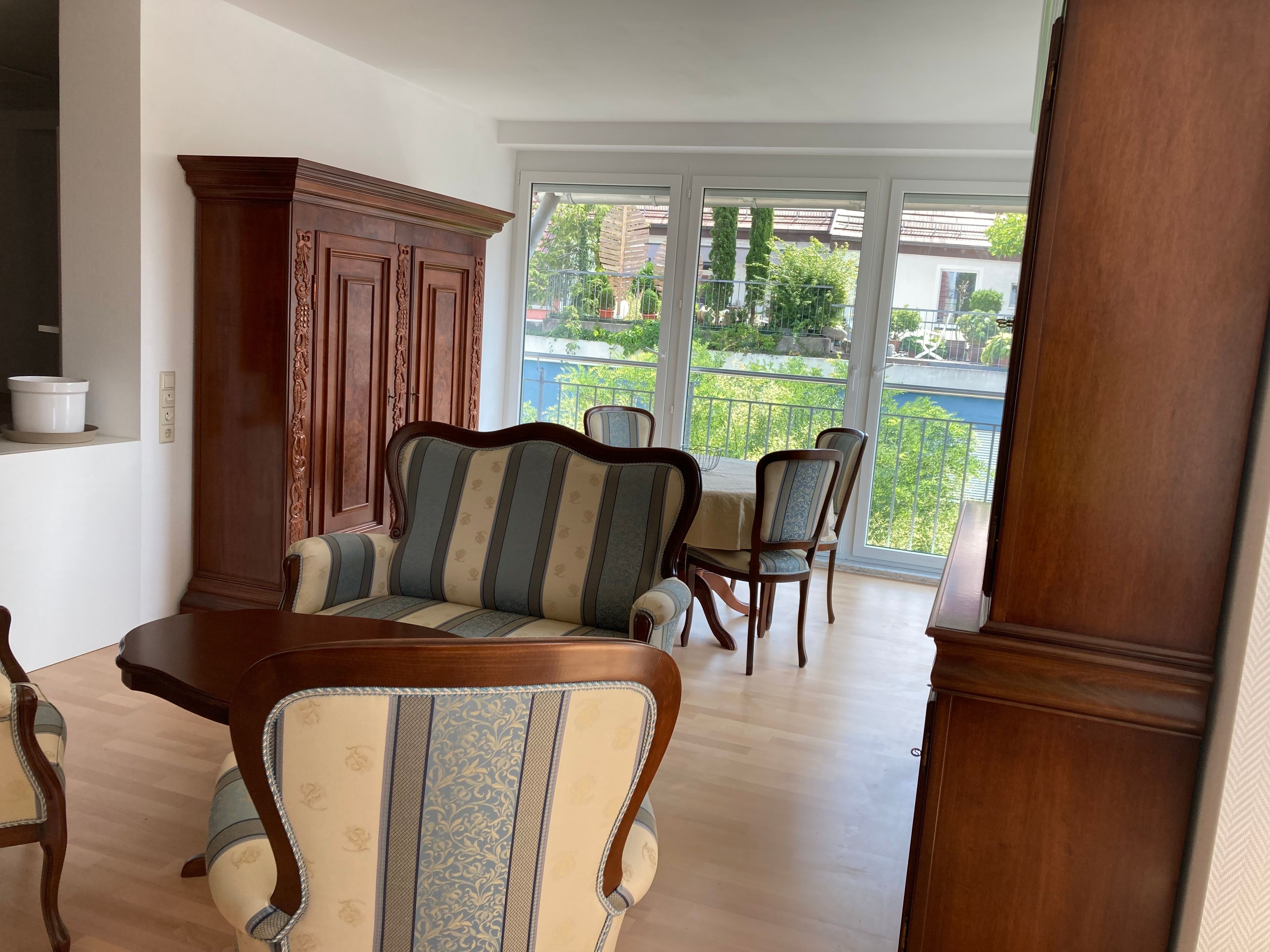 A dining area with a wooden table and chairs, a cabinet, and a view of a balcony with greenery.