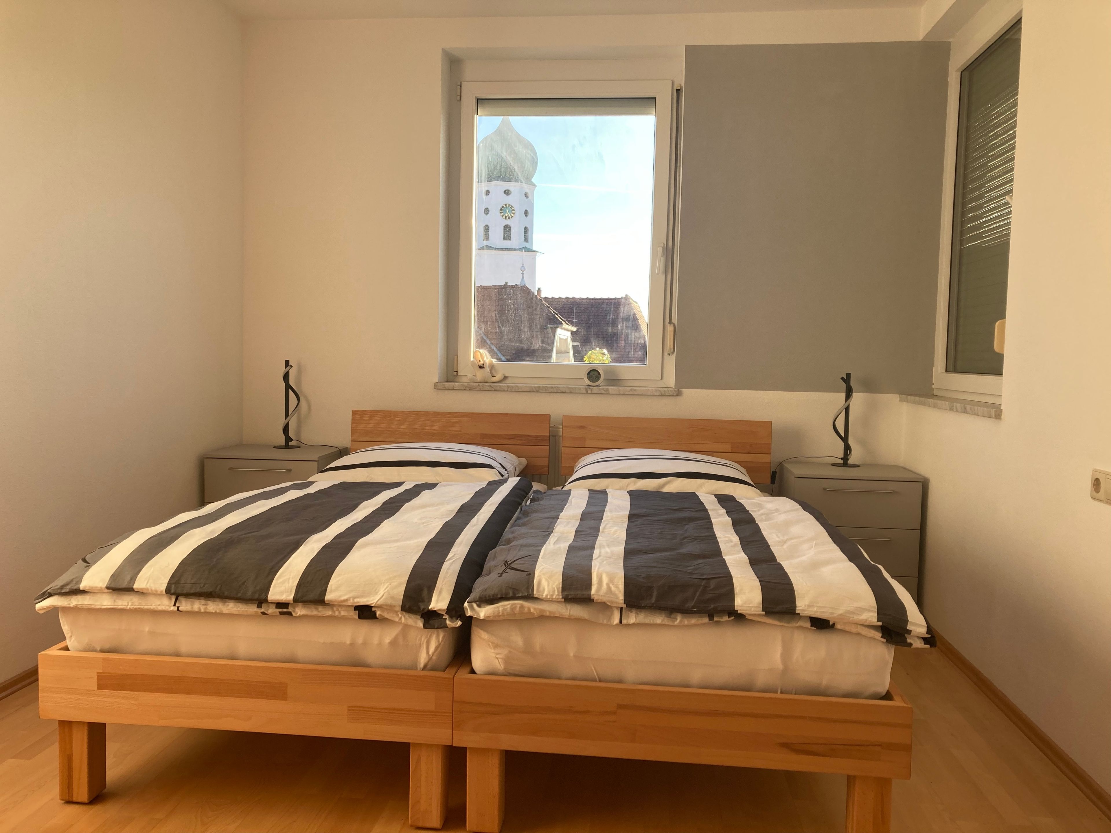 Ein Doppelbett mit gestreifter Bettwäsche, zwei Nachttische, ein Fenster mit Blick auf ein Gebäude und eine an der Wand befestigte Uhr.