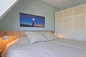 A bedroom with a bed, a nightstand, a wall-mounted lamp, a painting of a lighthouse, and a closet with louvered doors. - Residence de l'Europe / Noordstraat 19D (Domburg)