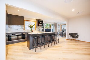 A modern kitchen with a central island, bar stools, and a dining area with chairs. - Hokowhitu Haven | Style, Space & Luxury (Palmerston North)