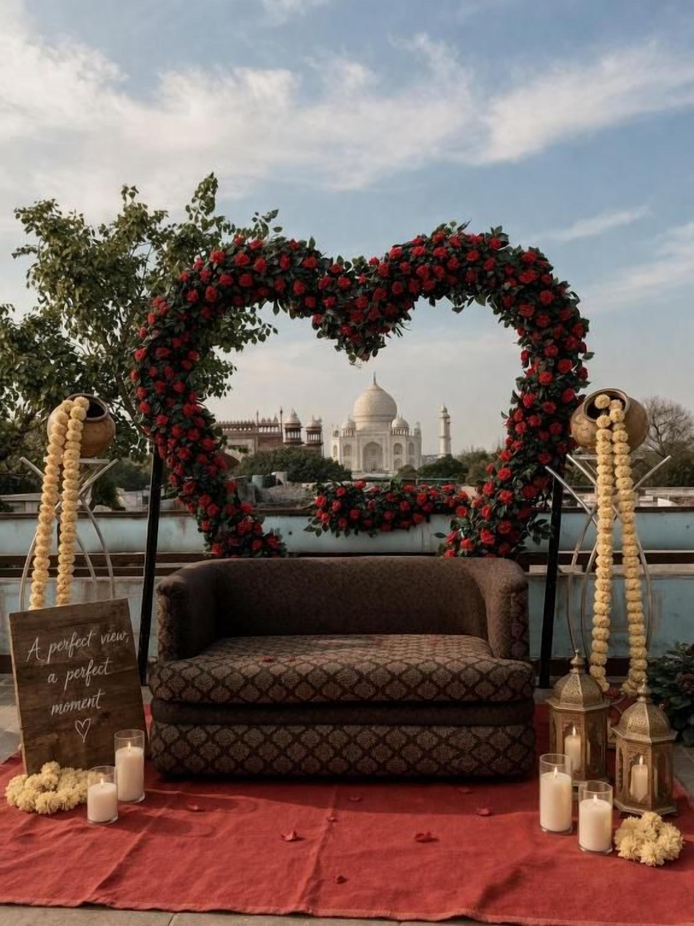 A heart-shaped floral arch with a sofa and candles, overlooking a body of water with a historic building in the background.