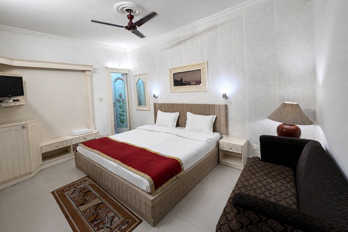 A hotel room with a bed, bedside tables, a TV, a ceiling fan, a lamp, and a patterned rug.
