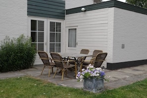 Un bâtiment blanc surmonté d’un toit végétalisé, une terrasse avec une table et des chaises, et un jardinier avec des fleurs.
