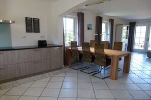 A dining area with a wooden table and chairs, a kitchen counter, and a window with curtains. - Holiday home with 2 bedrooms (Callantsoog)