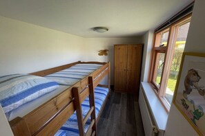 Een kleine kamer met een stapelbed, een houten garderobe en een raam met uitzicht op groen.
