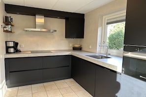 A modern kitchen with dark cabinetry, a stainless steel range hood, and a window providing natural light. - Holiday home Duynopgangh 27 (Julianadorp)