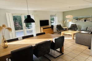 Een moderne woonkamer met een houten eettafel, zwarte stoelen, een bank, een televisie en een groot raam met luiken.
