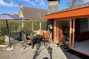 Een terras buiten met een tafel en stoelen, een overdekte zithoek en uitzicht op een huis met schoorsteen.
