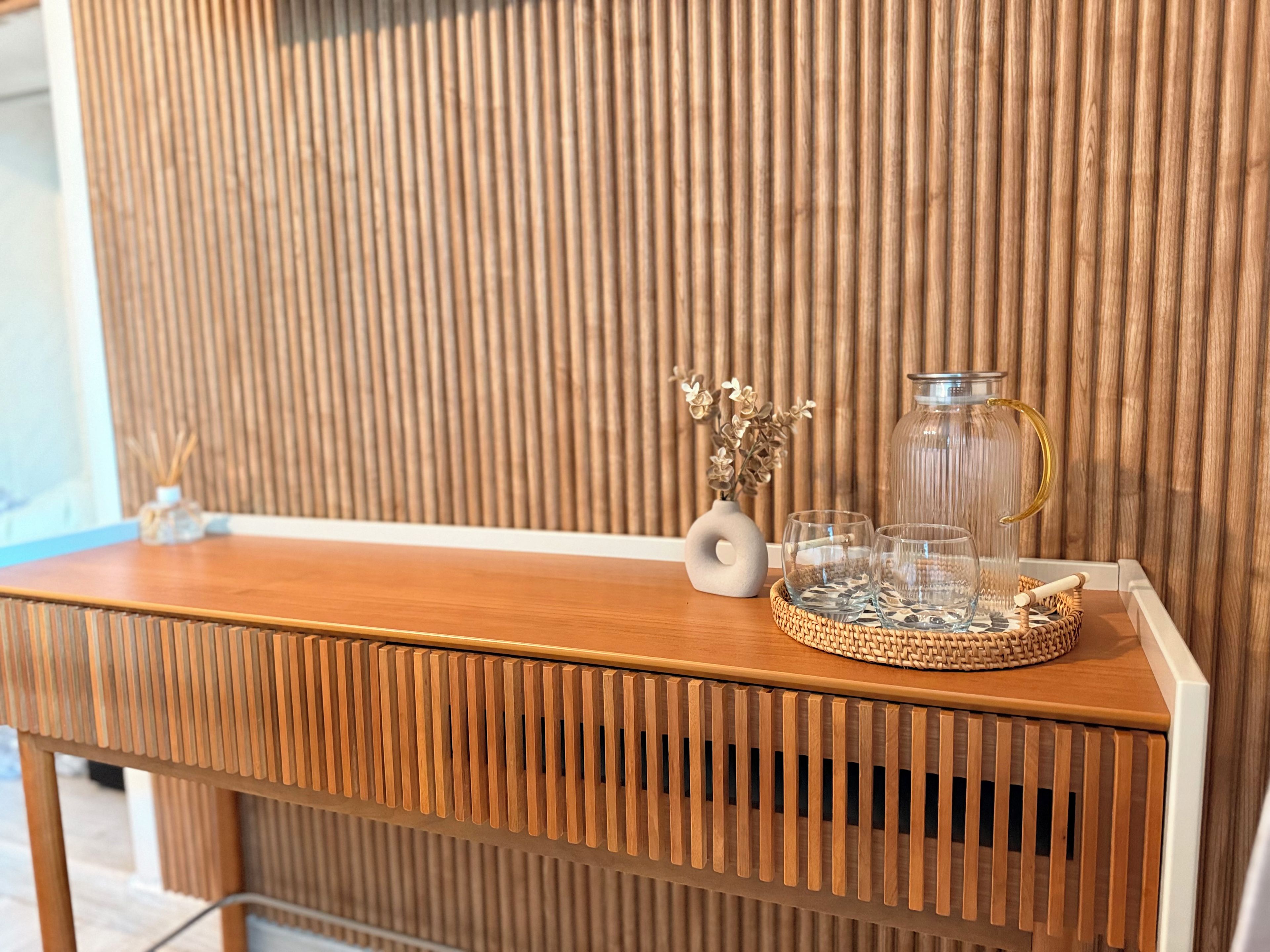 Um console de madeira com uma bandeja de fibra natural, um jarro de vidro e um vaso com flores secas.
