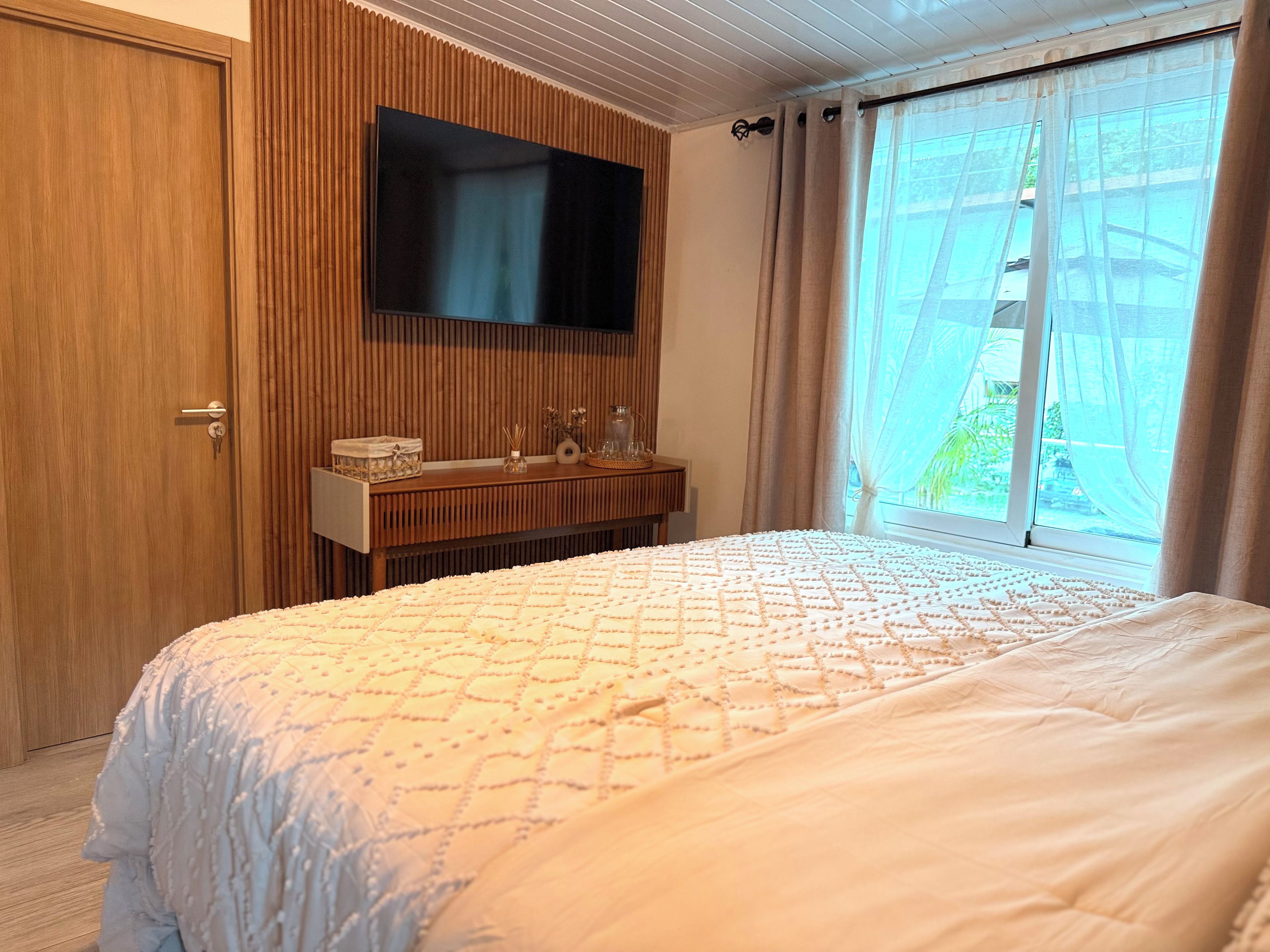 A bedroom with a bed, a TV mounted on the wall, a bedside table with a lamp, and a window with curtains.