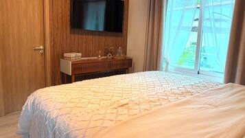 A bedroom with a bed, a TV mounted on the wall, a bedside table with a lamp, and a window with curtains.