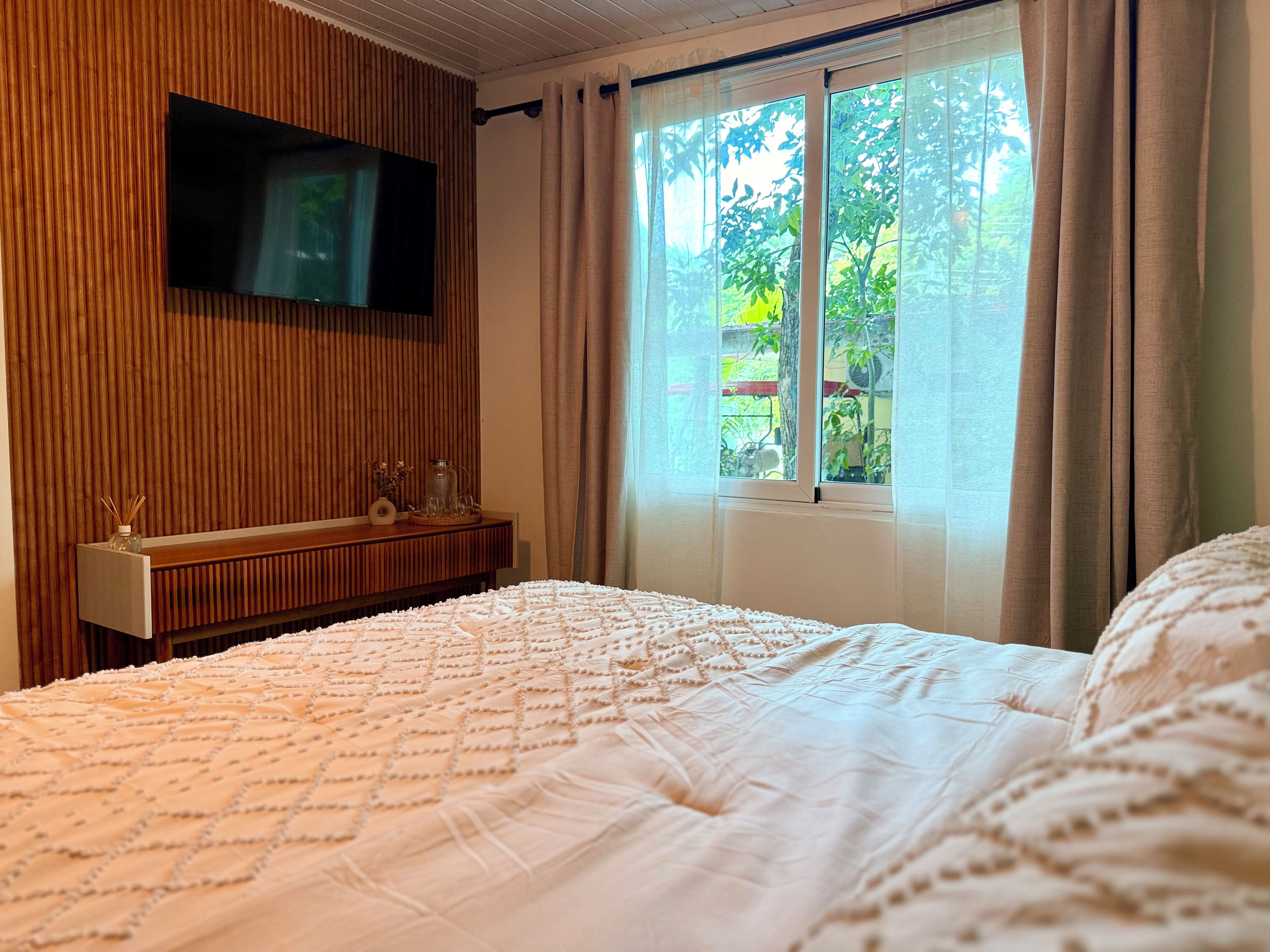 Une chambre à coucher avec un lit, un téléviseur fixé au mur, une fenêtre avec des rideaux et un mur recouvert de panneaux de bois.
