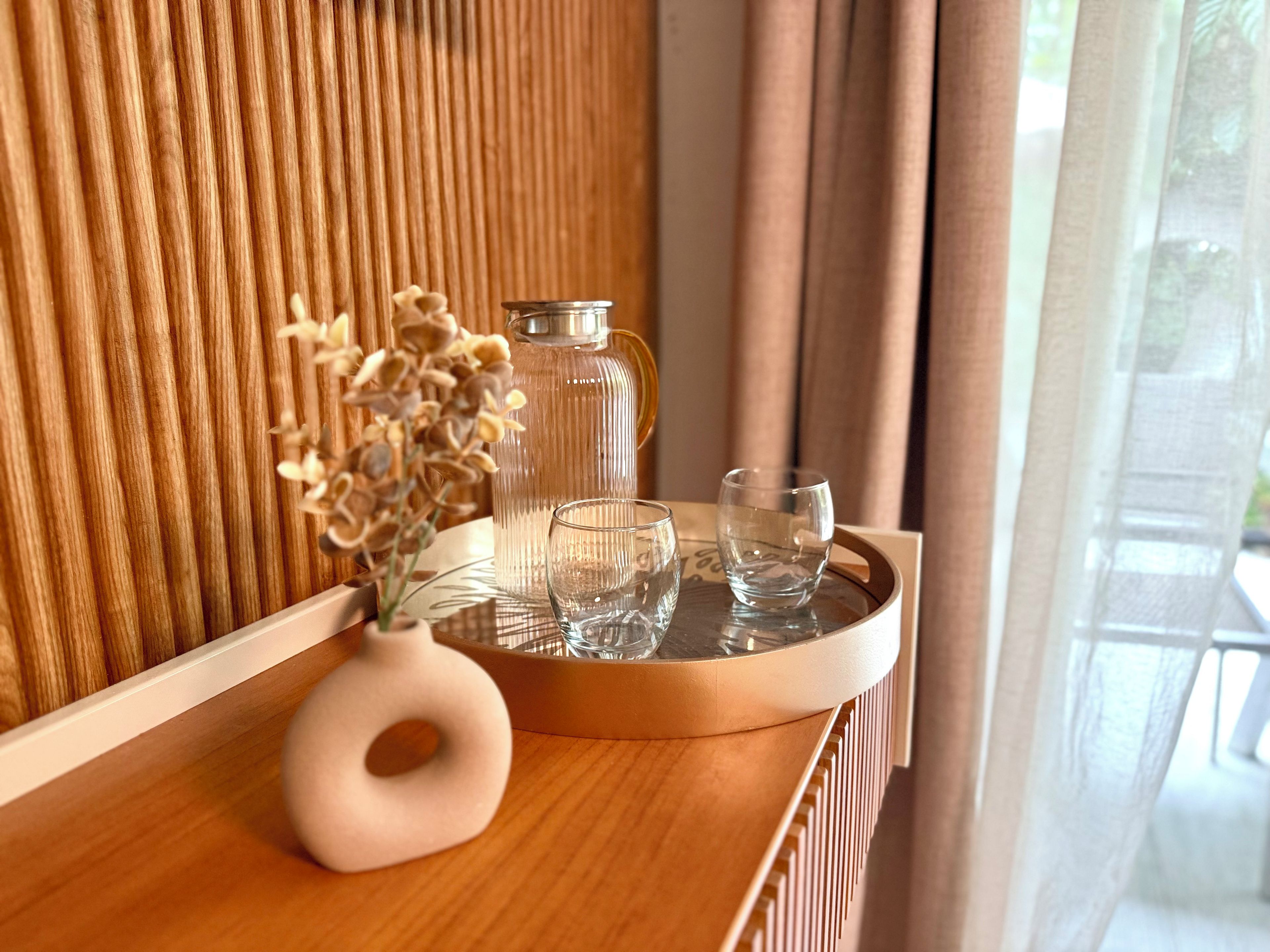 Un plateau en bois avec un vase et deux verres posés sur une table, une carafe en verre et un mur en bois orné de lignes verticales.