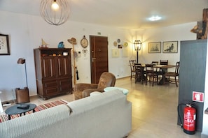 A living room with a sofa, armchair, and a dining area with a table and chairs. There is a wooden cabinet and a fire extinguisher visible. - Spacious apartment for 2, near Baleal Beach (Ferrel, Peniche)