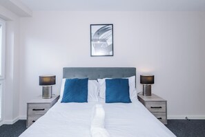 A neatly made bed with a white duvet and two blue pillows, flanked by two bedside tables with lamps, against a plain wall with a framed artwork above. - Spacious 2BDR with Modern Comfort Sleeps 4 (Manchester)
