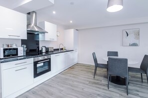 A modern kitchen with white cabinetry, stainless steel appliances, and a dining area with a round table and grey chairs. - Spacious 2BDR with Modern Comfort Sleeps 4 (Manchester)