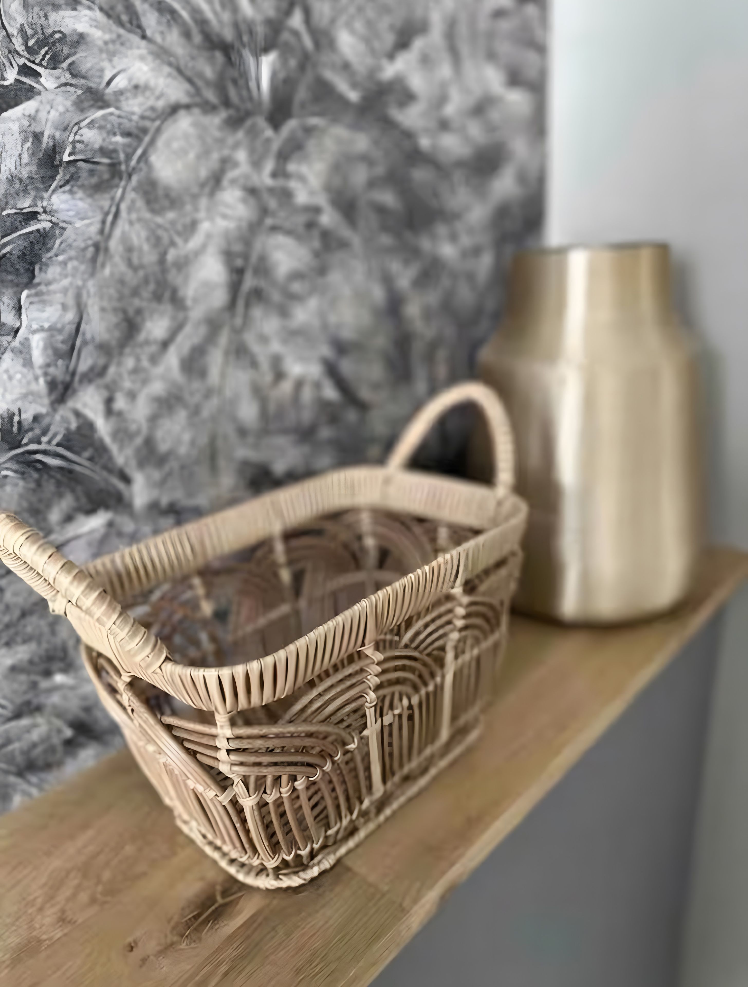 A wicker basket on a wooden shelf with a textured wall and a metallic vase in the background.