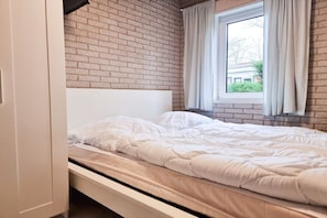 Een enkel bed met een wit hoofdeinde en beddengoed, een raam met gordijnen en een bakstenen muur.
