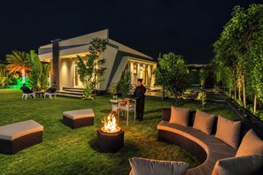 Een terras buiten met een vuurkuil, loungesessels en een modern gebouw op de achtergrond.