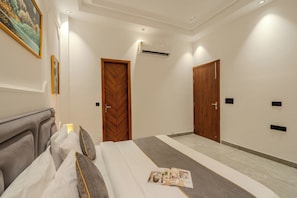 A hotel room with a bed, a bedside table, a nightstand, a wall-mounted air conditioner, and a door. - ELIVAAS Signature Aarisha 3BHK Villa (New Delhi)