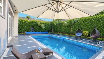 A swimming pool with lounge chairs and a table, surrounded by a covered area with a view of a green hedge and a building.