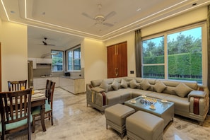 A modern living room with a sofa, ottomans, a glass coffee table, and a dining area with chairs and a table. - ELIVAAS Signature Aarisha 3BHK Villa (New Delhi)