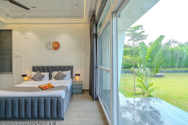 A modern bedroom with a large bed, bedside lamps, a wall clock, and a view of a garden through a sliding door. - ELIVAAS Signature Aarisha 3BHK Villa (New Delhi)