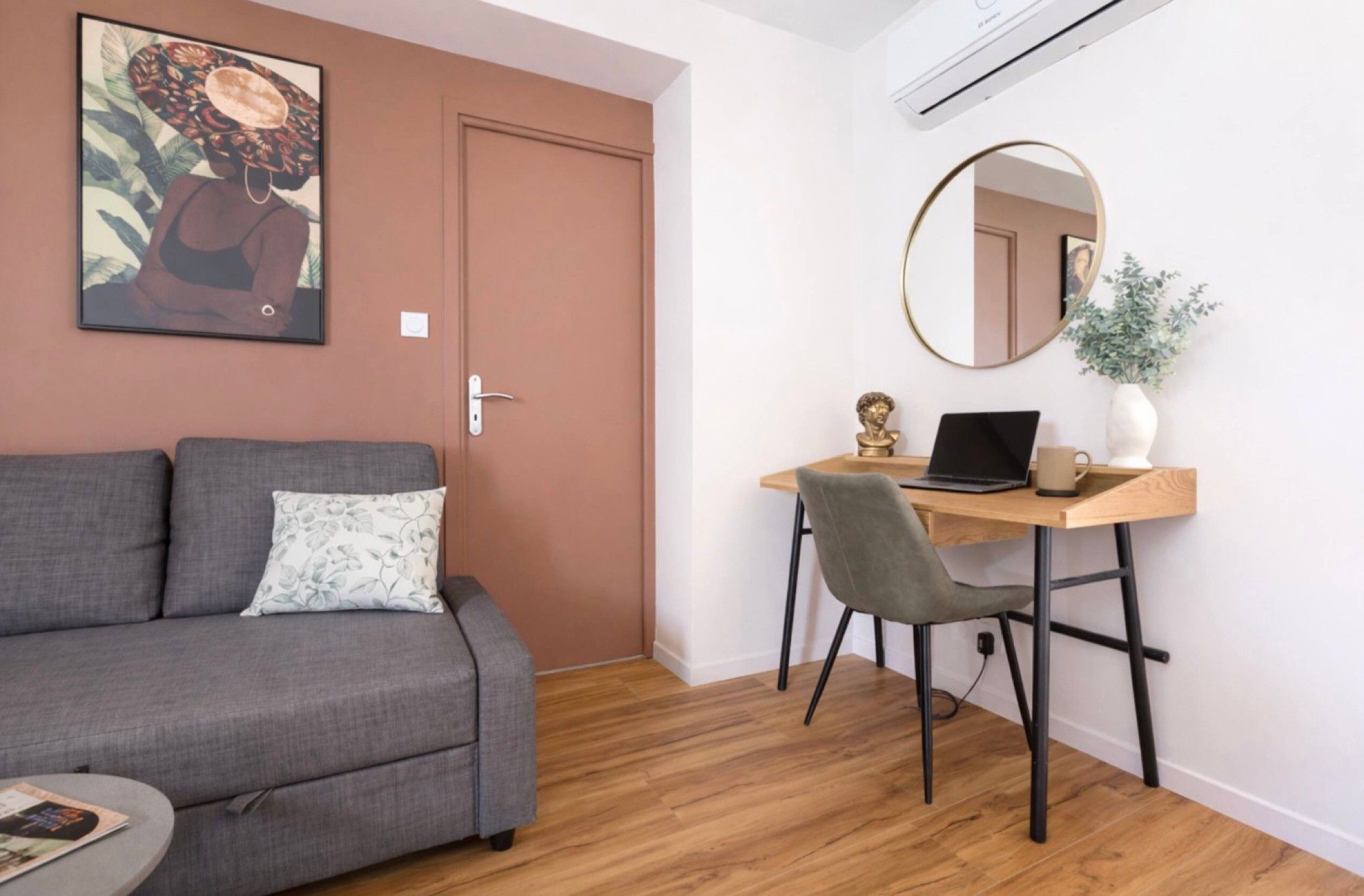 A room with a grey sofa, a wooden desk, a laptop, a grey chair, a round mirror, and a potted plant.