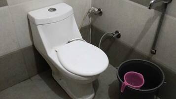 A toilet with a bidet attachment, a black bucket with a pink scoop, and a wall with a checkered pattern.