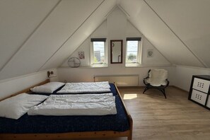 A room with a sloped ceiling, two windows, a bed with white bedding, a chair with a fur throw, and a radiator. - Beach Parcel 207 in the Strandslag holiday park (Julianadorp)
