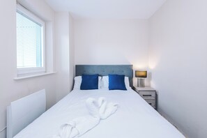 A neatly made bed with white linens and two blue pillows, a bedside table with a lamp, and a window with blinds. - Bright and Modern 2BDR 2BA Relaxing City Stay (Manchester)