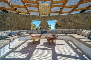 A sunlit outdoor seating area with stone walls, a wooden pergola, and a view of the sky. - Unparalleled luxury and warmth meet sweeping Aegean blues - The ultimate Mykonos retreat (Mykonos)