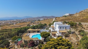 A white building with a pool, surrounded by greenery and a view of a coastal town. - Unparalleled luxury and warmth meet sweeping Aegean blues - The ultimate Mykonos retreat (Mykonos)