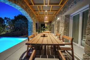 Un área de comedor al aire libre con piscina, paredes de piedra y mobiliario de madera.