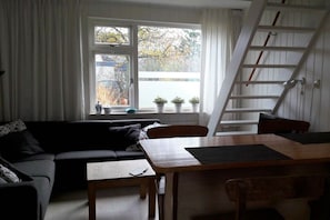 A living room with a sofa, a dining table, and a window with potted plants. - Holiday apartment with 2 bedrooms (Callantsoog)