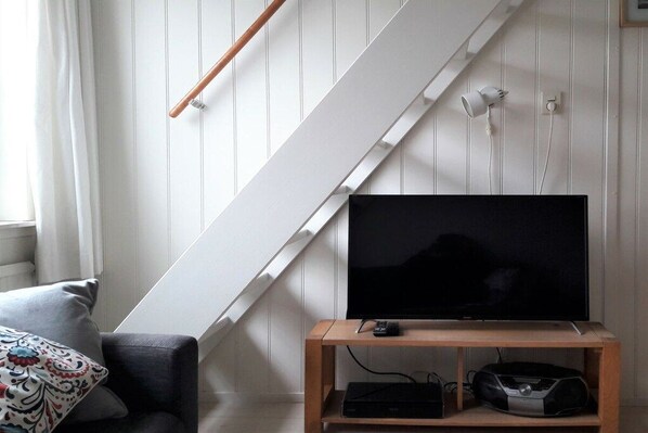 A living room with a flat-screen TV on a wooden stand, a sofa with patterned cushions, and a white staircase. - Holiday apartment with 2 bedrooms (Callantsoog)