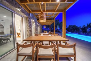 An outdoor dining area with a wooden table and chairs, a pergola overhead, and a view of a swimming pool at dusk. - Secluded Cycladic serenity meets an endless blue horizon in Mykonos (Mykonos)