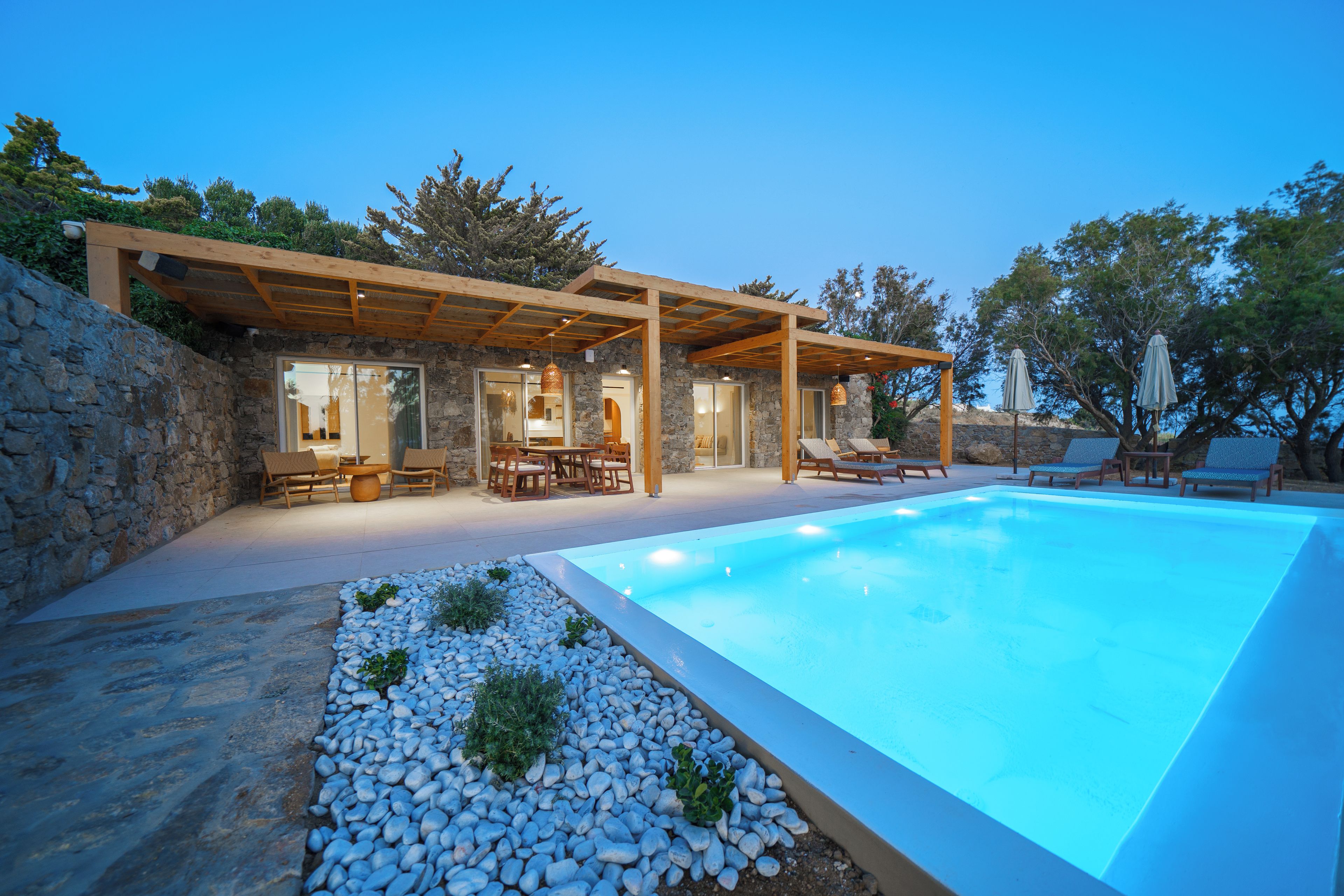 A modern outdoor pool area with a stone wall, wooden pergola, and lounge chairs.
