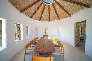 A dining area with a round wooden table and chairs, a thatched roof ceiling, stone walls, and windows with views of greenery. - Unrivaled grandeur and absolute seclusion meet infinite Aegean horizons (Mykonos)