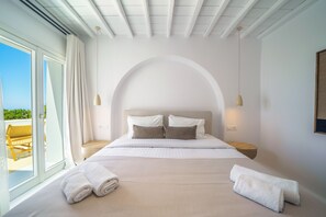 A bedroom with a large bed, a view of the outdoors, and a wooden side table. - Unrivaled grandeur and absolute seclusion meet infinite Aegean horizons (Mykonos)