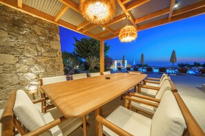 An outdoor dining area with a long wooden table and chairs, a stone wall, and a view of the sea. - Unrivaled grandeur and absolute seclusion meet infinite Aegean horizons (Mykonos)
